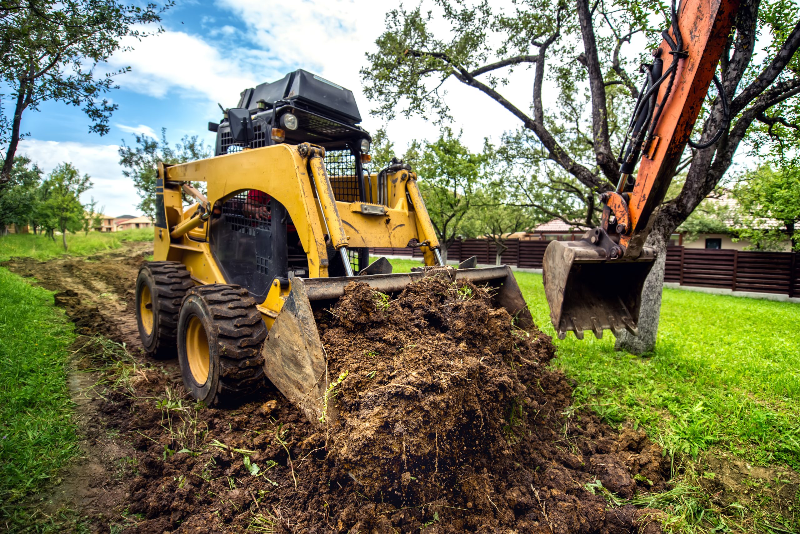 A skid steer loader moving soil next to an excavator on a green property, showing the groundwork and preparation phase of a hardscape project.