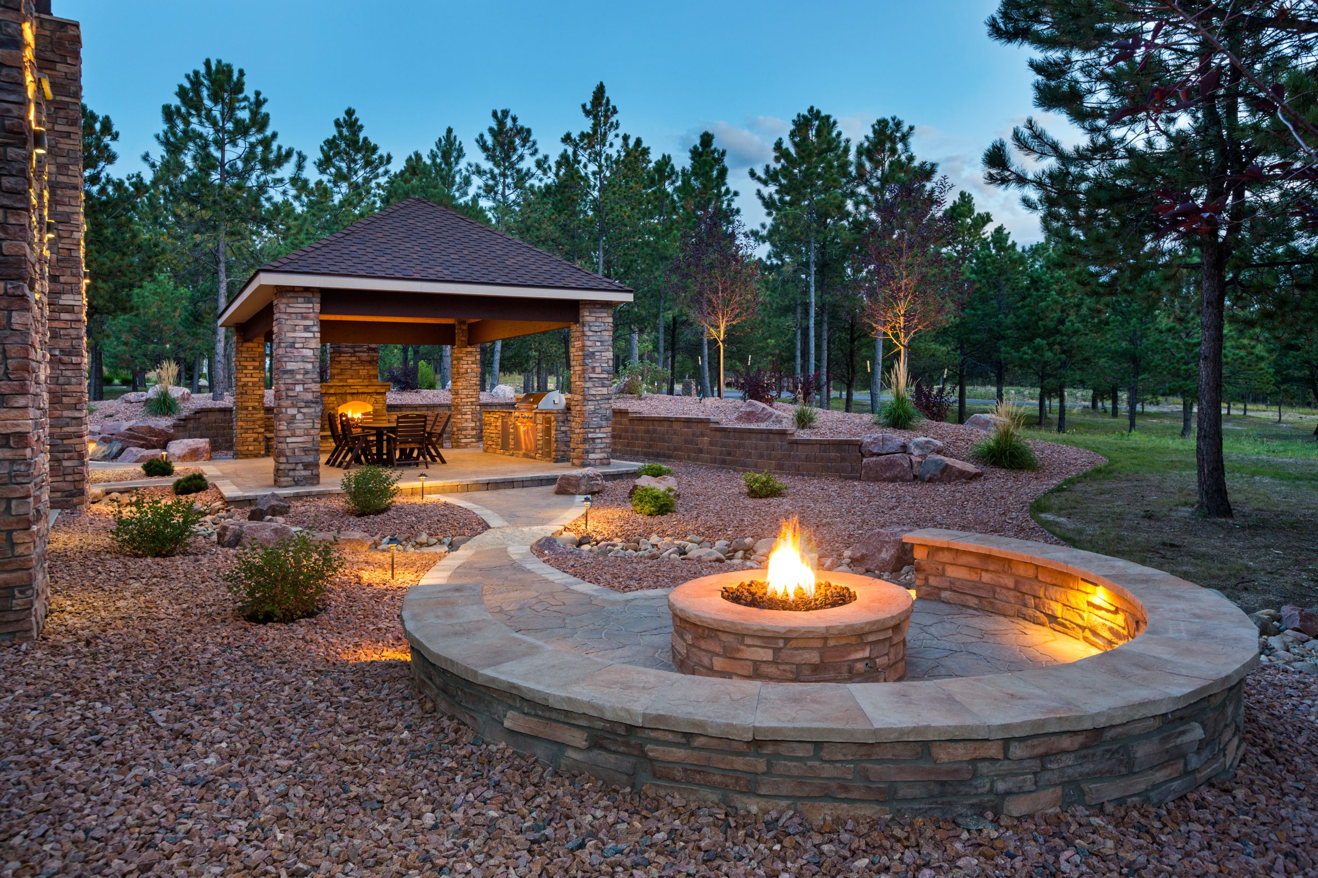 A beautifully designed backyard featuring a stone patio with a circular fire pit, soft landscape lighting, and a covered outdoor dining pavilion surrounded by pine trees at dusk. Perfect example of professional hardscape design and craftsmanship.