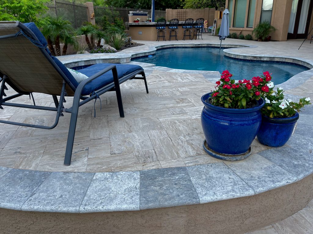 A travertine tiled pool deck with a spa and outdoor kitchen on a desert landscaped backyard in Arizona.